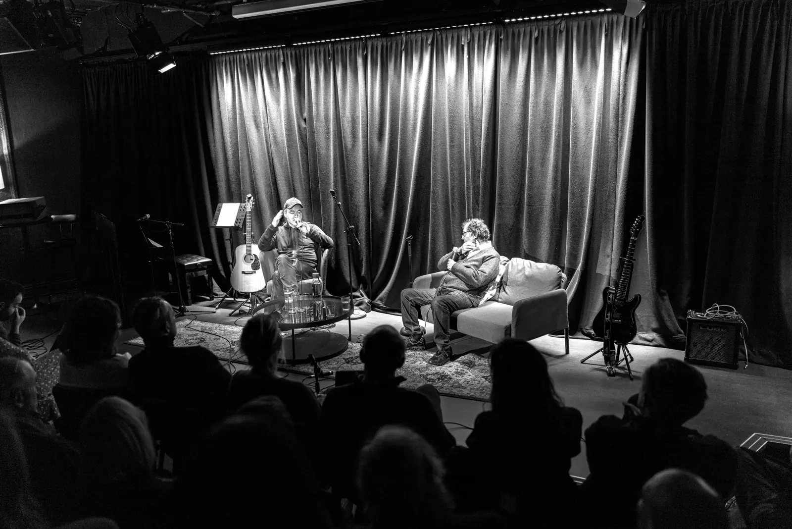 The Circle & Star stage set for a salon — two performers in armchairs beneath the house curtain, the audience in silhouette across the front of frame.
